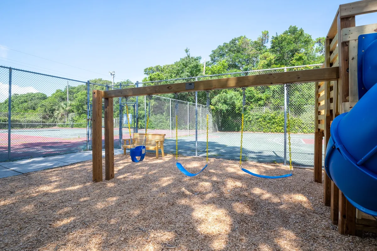 A cheerful recreation area featuring swings, a slide, a bench for parents or caregivers, and adjacent tennis courts. Children are seen playing, while families enjoy both the playground and the opportunity for a friendly tennis match nearby.