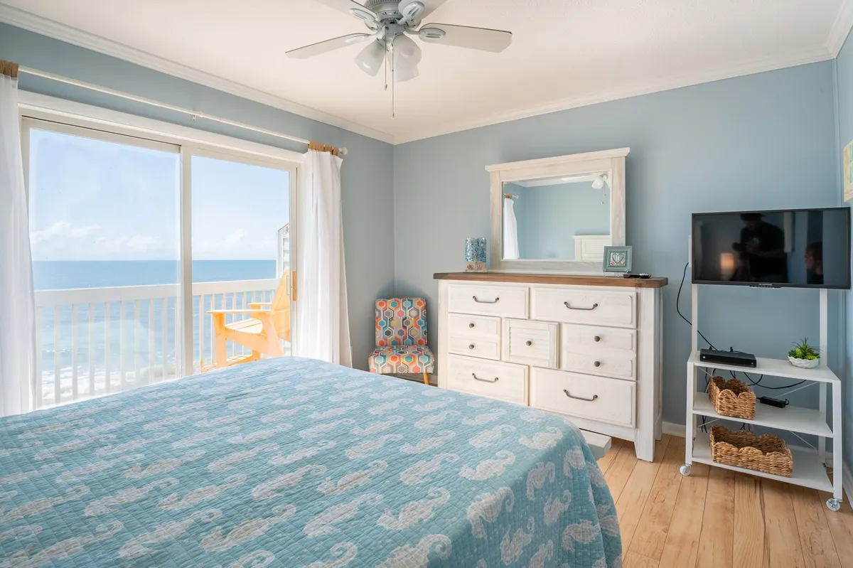 A serene primary bedroom with a bed dressed in a blue quilt, a large window framing sweeping ocean views, a dresser, and a colorful accent chair. The soft natural light and tranquil color palette create an inviting, restful atmosphere ideal for a private retreat.