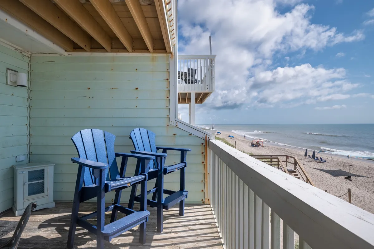 A covered oceanfront deck furnished with blue Adirondack chairs, overlooking the sandy beach and gently rolling waves. The tranquil setting is perfect for sipping morning coffee, enjoying the sea breeze, or gathering as the sun rises over the ocean.