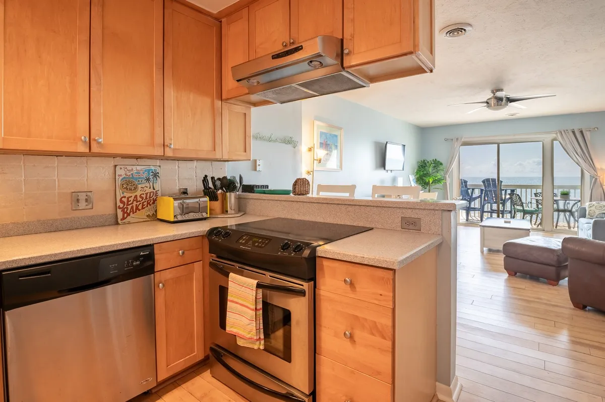 A view of the kitchen with modern appliances, warm cabinetry, and the dining area visible in the background. The open layout encourages cooking, casual conversation, and shared meals, with sunlight streaming through nearby windows enhancing the welcoming atmosphere.