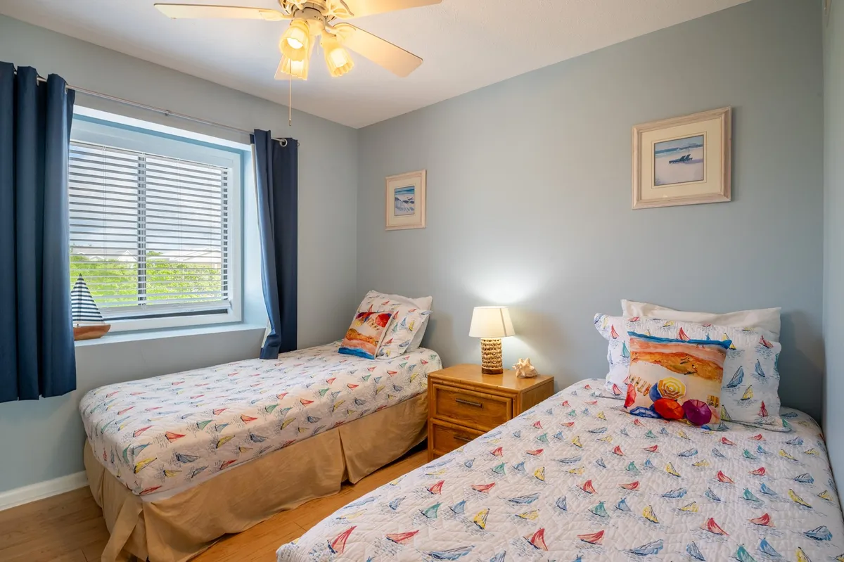 A light and cheerful second bedroom featuring two twin beds with colorful bedding, a window with sheer curtains allowing natural light to brighten the space, and a cozy nightstand with a lamp between the beds. The room exudes a welcoming, playful vibe perfect for kids or additional guests.