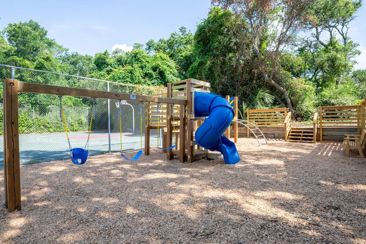 A cheerful, well-equipped playground featuring a swing set, a blue slide, and a climbing structure. The space is clearly designed for kids, with bright colors and spacious grounds for running and imaginative play.