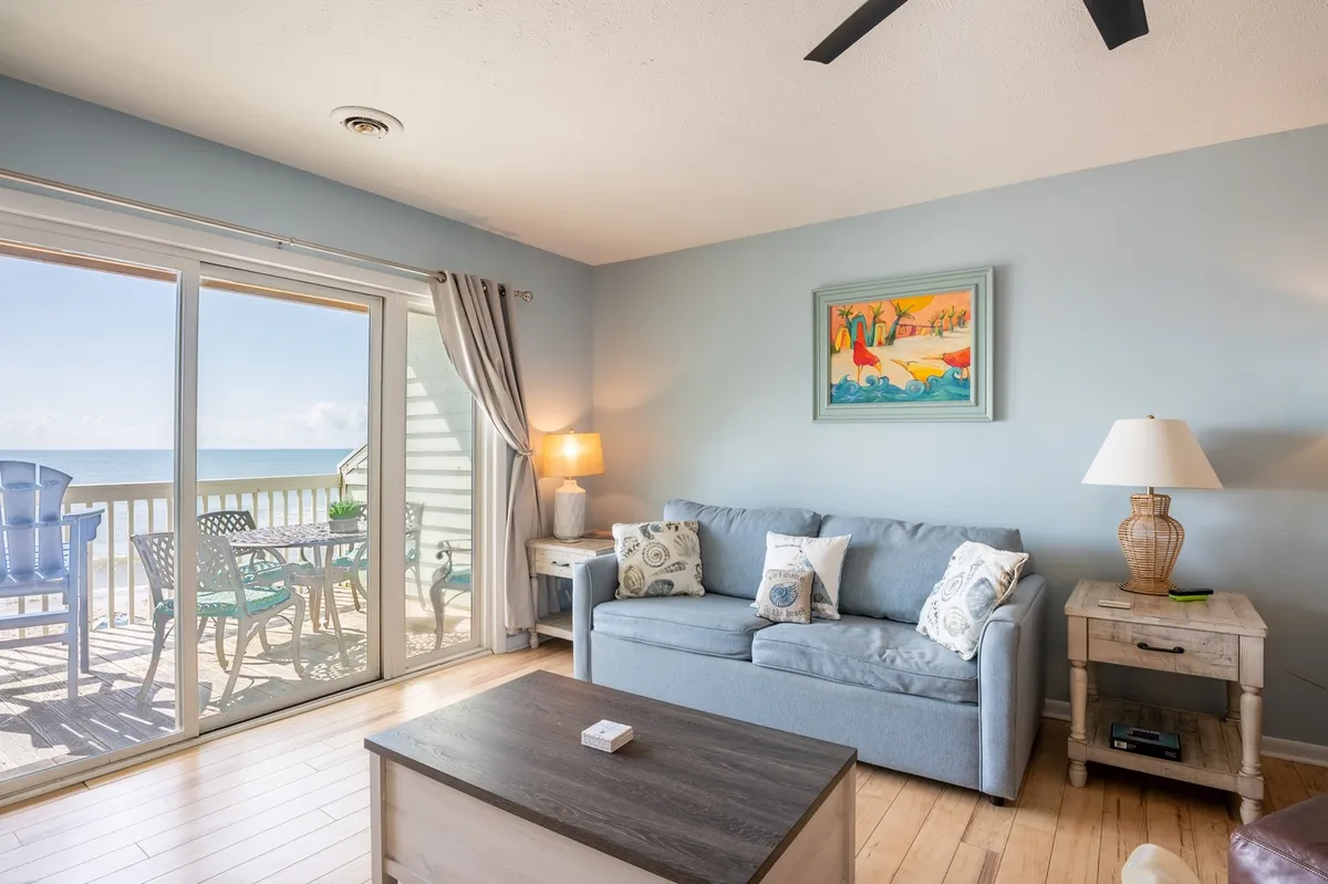 A bright, open living room featuring a plush sofa set near large sliding glass doors that open onto an oceanfront deck. Tasteful artwork, soft lighting from lamps, and a well-placed coffee table create a welcoming space for relaxation and socializing. Through the glass doors, you can glimpse the ocean, bringing in natural light and a coastal atmosphere.