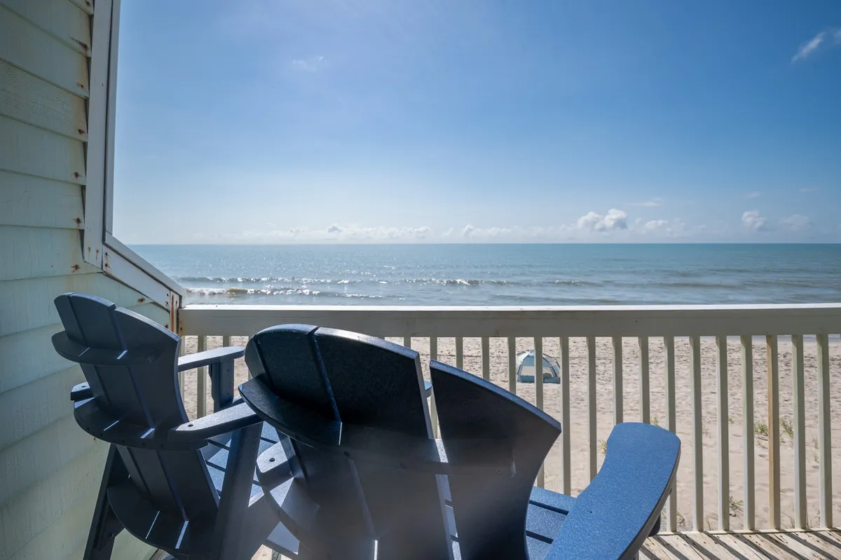 This hero image captures a tranquil view from the condo's private deck, featuring inviting adirondack chairs facing a stunning expanse of ocean waves, soft sandy beach, and an open sky adorned with gentle clouds. The scene radiates relaxation, with the promise of serene mornings and peaceful evenings spent soaking in the sights and sounds of the Atlantic coastline.