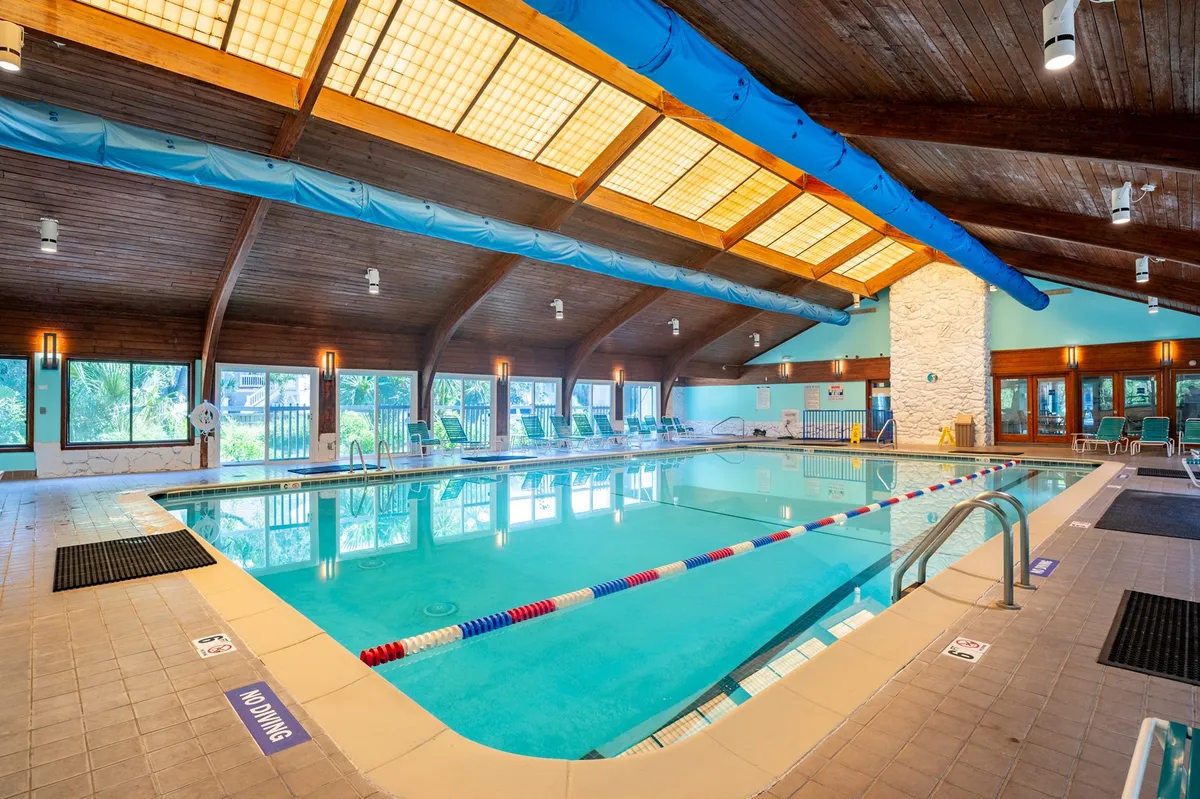 An inviting indoor swimming pool with a high wooden ceiling, surrounded by lounge chairs. Sunlight streams in through large windows, creating a cheerful atmosphere, and the pool area is designed for both relaxation and fun for all ages.