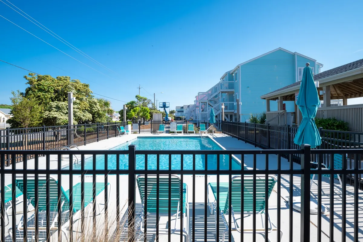 A vibrant outdoor pool area featuring sparkling blue water, lounge chairs, umbrellas, and lush greenery. Guests are seen swimming, sunbathing, and socializing, capturing the lively seasonal pool scene.